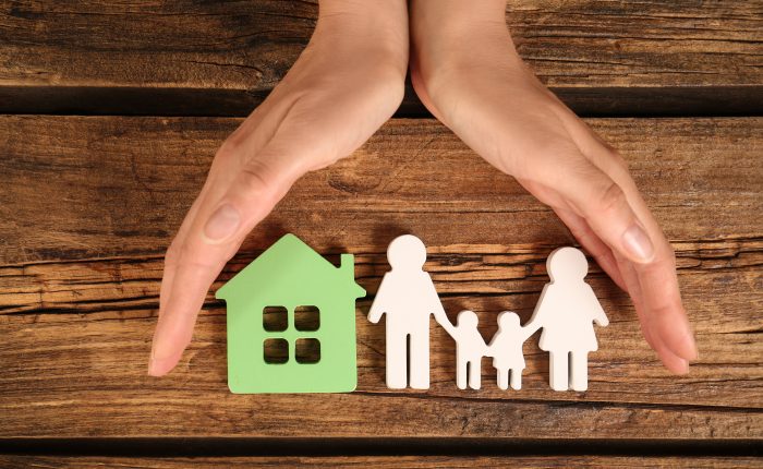 Woman holding hands near figures of house and family on wooden background, top view